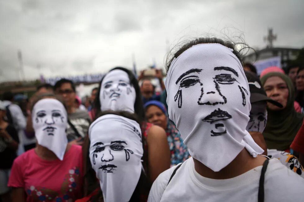 In front of a crowd are standing four persons. Each is wearing a white cloth, with a crying face drawn on it, over the head.